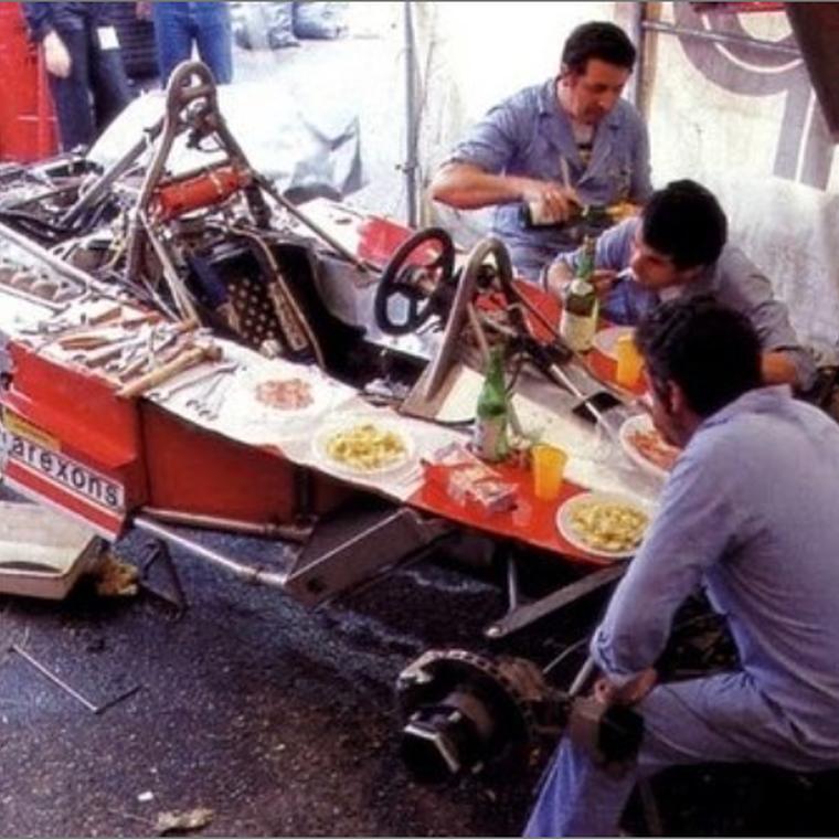 A racing team eating lunch beside a stripped-down car in the pit lane.